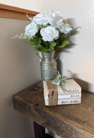 A corner in the house on the property with a set of books that reads "Shady Nook Est 2021" in front of some flowers